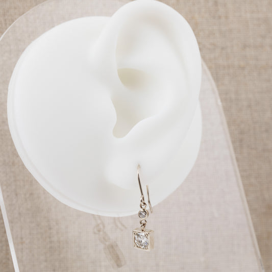A silver-toned dangle earring with a diamond displayed on a white silicone ear-shaped stand over a beige textured fabric background.