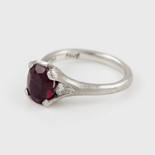 A platinum ring featuring a large deep red ruby gemstone with a textured band and side accent diamond, shown on a light gray background from an angle
