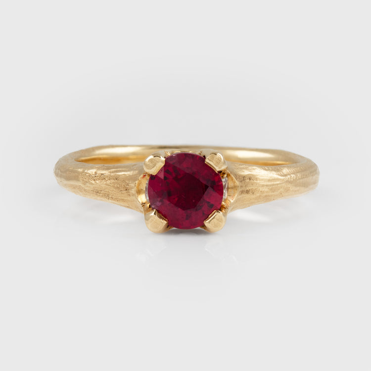 A gold ring with a textured band and a round-cut deep red gemstone in a prong setting, photographed on a light gray background.