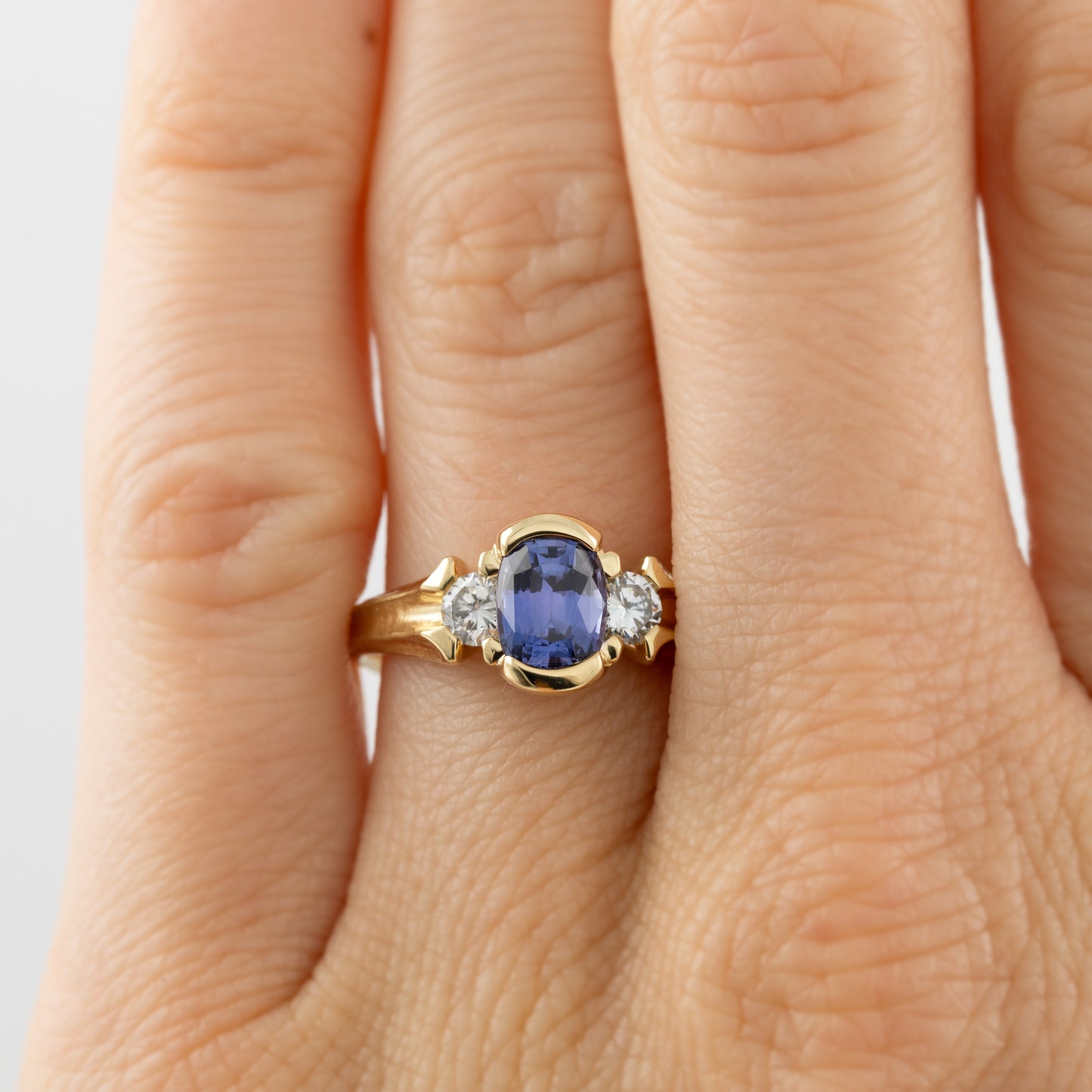 Yellow gold ring featuring half bezel set oval purple Sapphire flanked by round white Diamond accents pictured on a hand