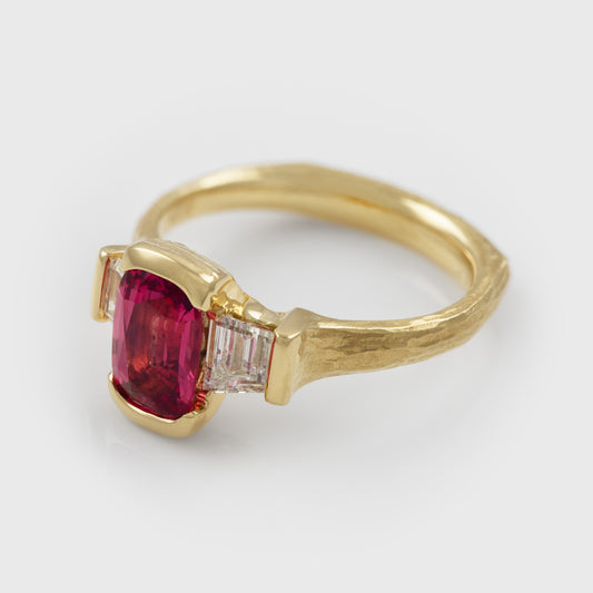 A yellow gold ring featuring a rectangular pinkish spinel center gemstone and two diamonds on either side, pictured at an angle and set against a light gray background.