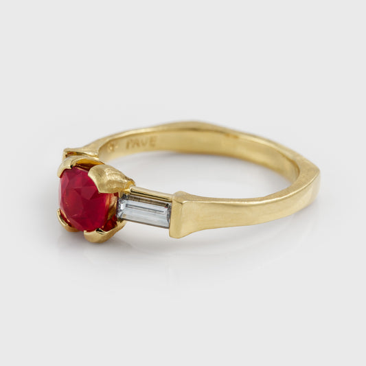 A yellow gold ring featuring a central red o pink spinel gemstone flanked by two diamonds, set against a light gray background at an angled view