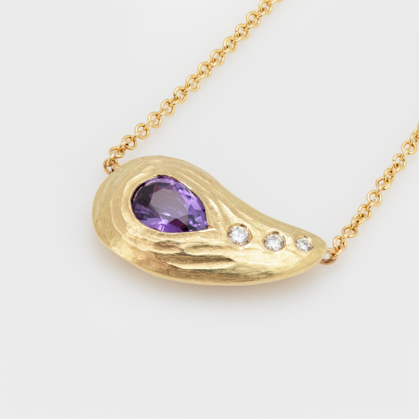 Close-up of a 18k yellow gold necklace featuring a pear shaped purple sapphire, and round brilliant cut diamonds.