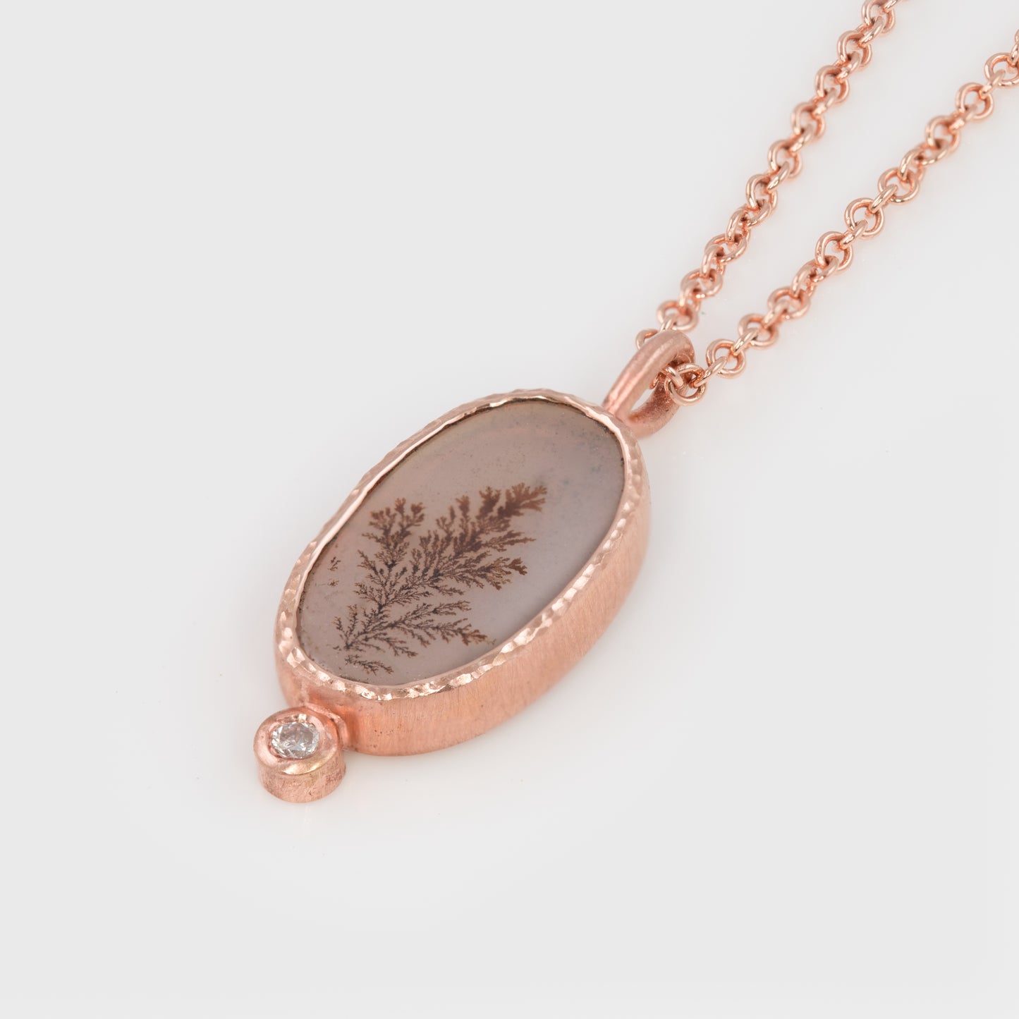 Close-up of a 14k rose gold necklace with an oval-shaped dendritic agate slice and a round brilliant-cut white diamond.