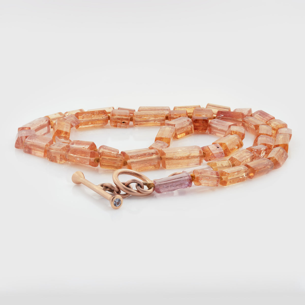 Beaded necklace featuring faceted topaz beads in varying shades of orange and pink, accented by lavender spinel and a rose gold toggle clasp, photographed on a white background.