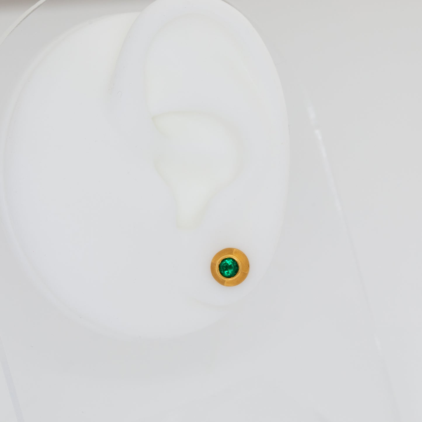 Close-up of a white ear mannequin with a green gemstone stud earring on a light gray background
