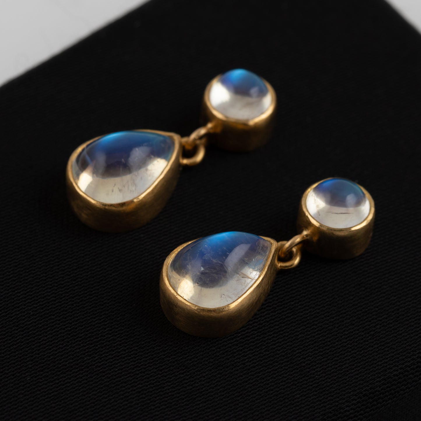 A pair of gold drop earrings with teardrop-shaped moonstone gemstones and round cabochon accents, displayed on a black surface.