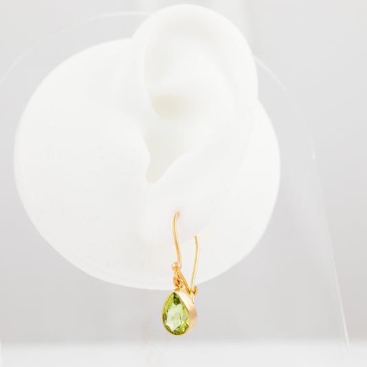 A gold dangle earring with a faceted green teardrop gemstone, displayed on a white ear-shaped jewelry stand against a light gray background.