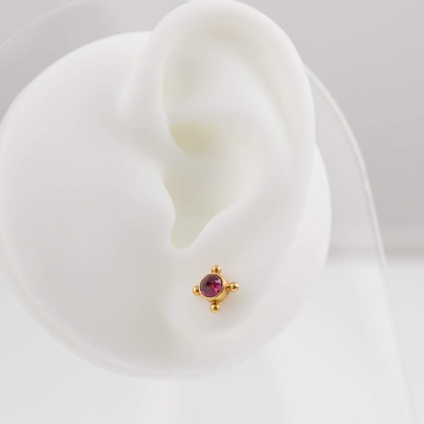 A small gold stud earring with a round pinkish-purple gemstone, displayed on a white silicone ear stand against a light gray background.
