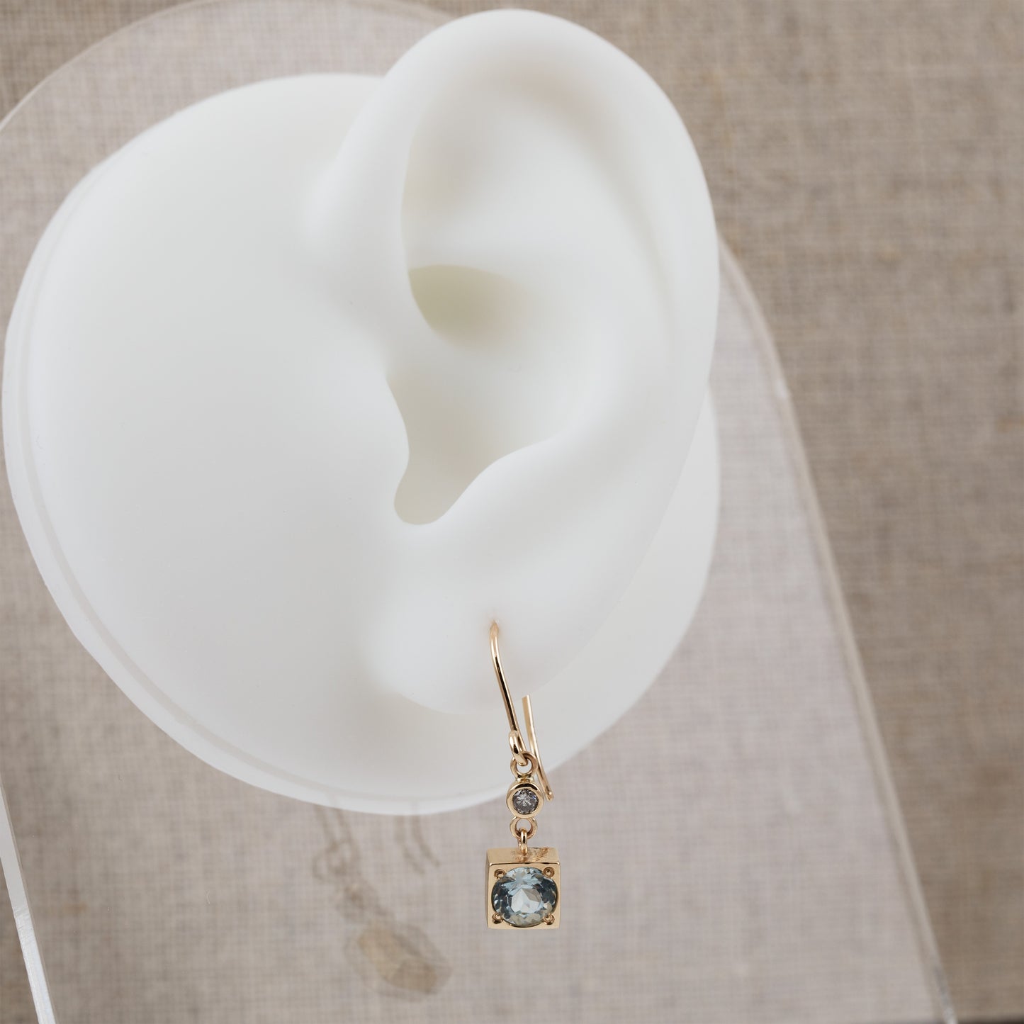 A gold drop earring in a square shape with a light blue aquamarine gemstone and a small round accent stone, displayed on a white silicone ear-shaped stand against a neutral beige textured background