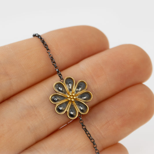 Gold and black flower-shaped pendant on a chain held between fingers against a white background