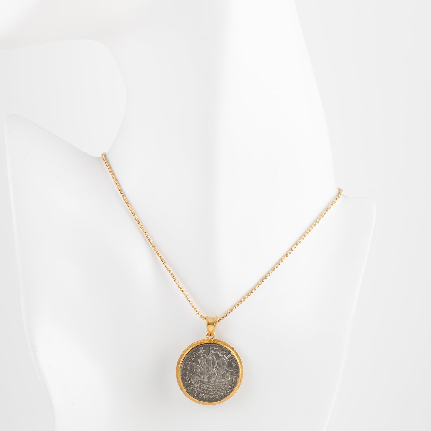 A gold chain necklace with a circular pendant featuring a vintage silver-toned coin, displayed on a white mannequin bust against a white background.