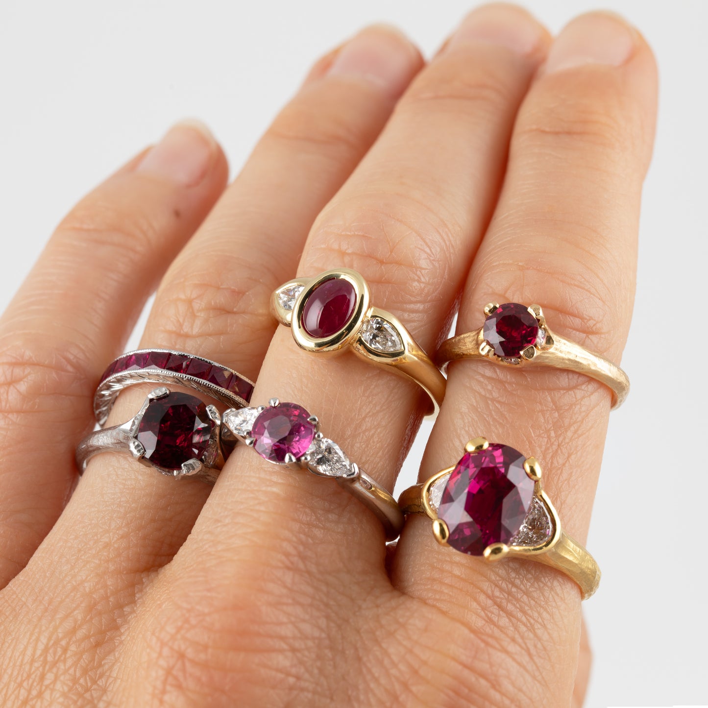 Six different rings featuring round and oval ruby gemstones in gold and platinum settings, displayed on a person's hand against a light gray background.