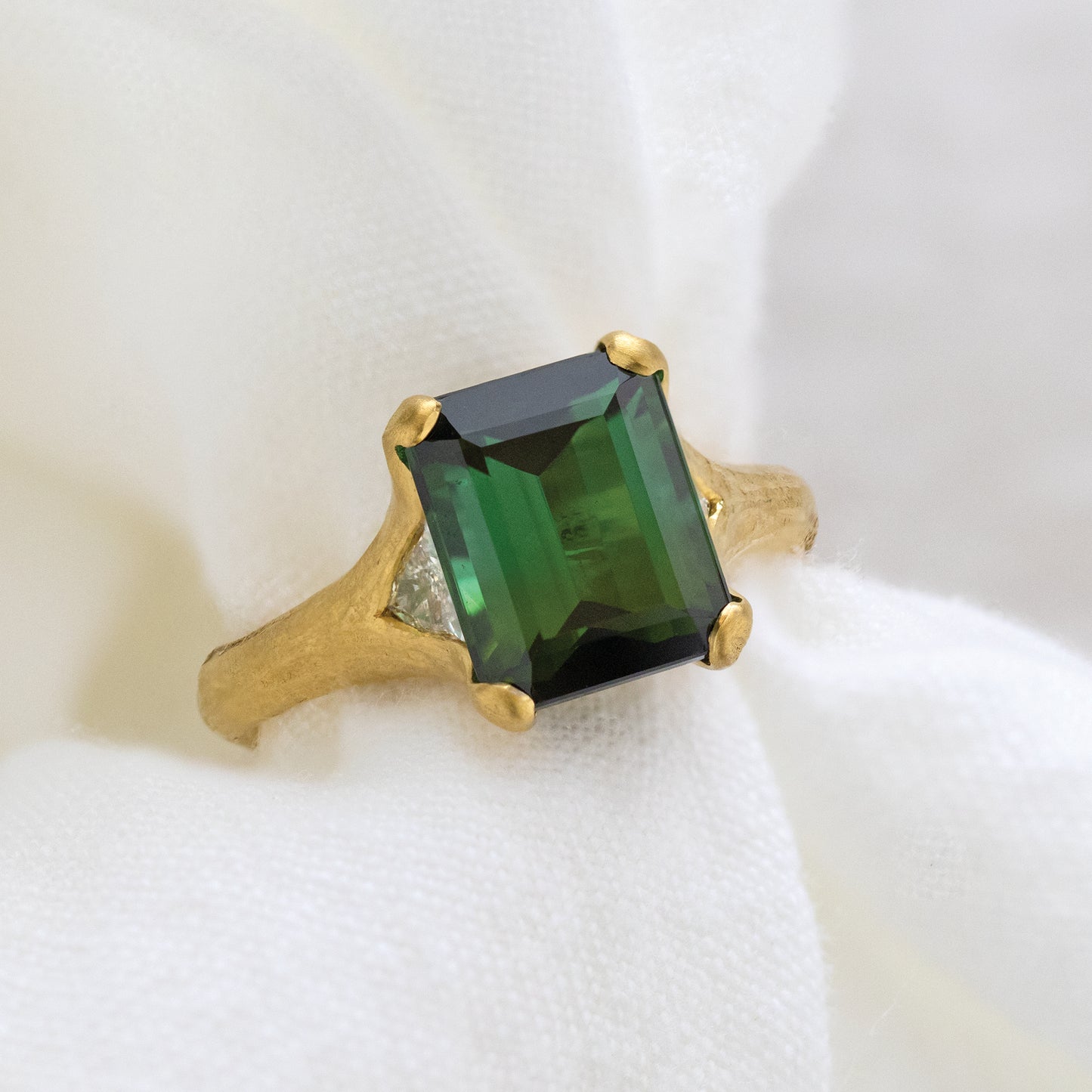 Close-up of a 22k yellow gold ring with natural gems, an emerald cut bicolor tourmaline, and 2 channel-set trillion cut diamonds.