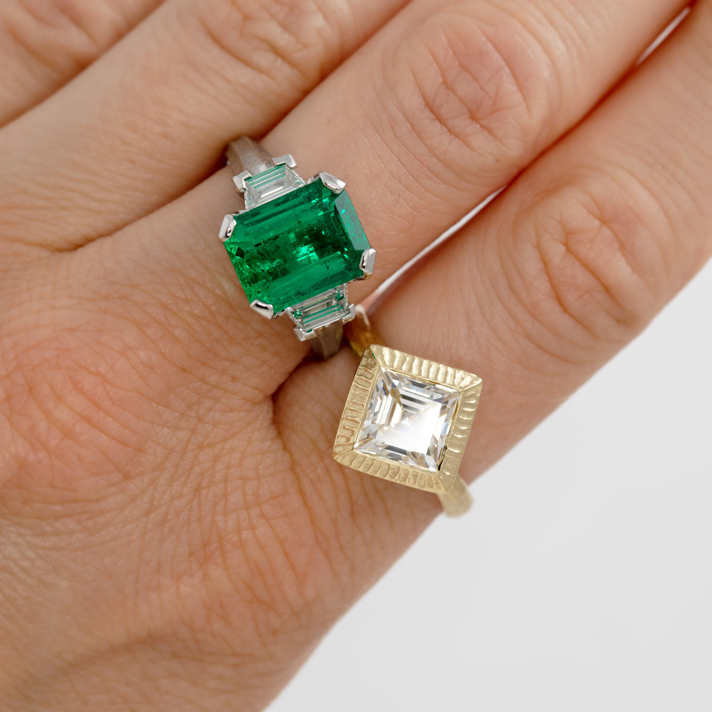 Two rings with emerald and diamond stones on a hand against a white background