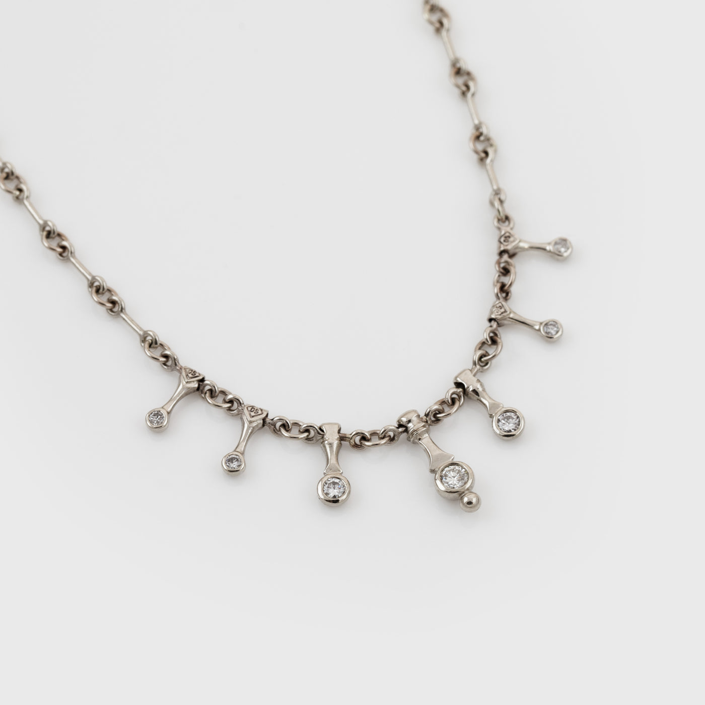 A silver-toned necklace with a chain and seven small dangling pendant charms, each set with a diamond, displayed on a light gray background.