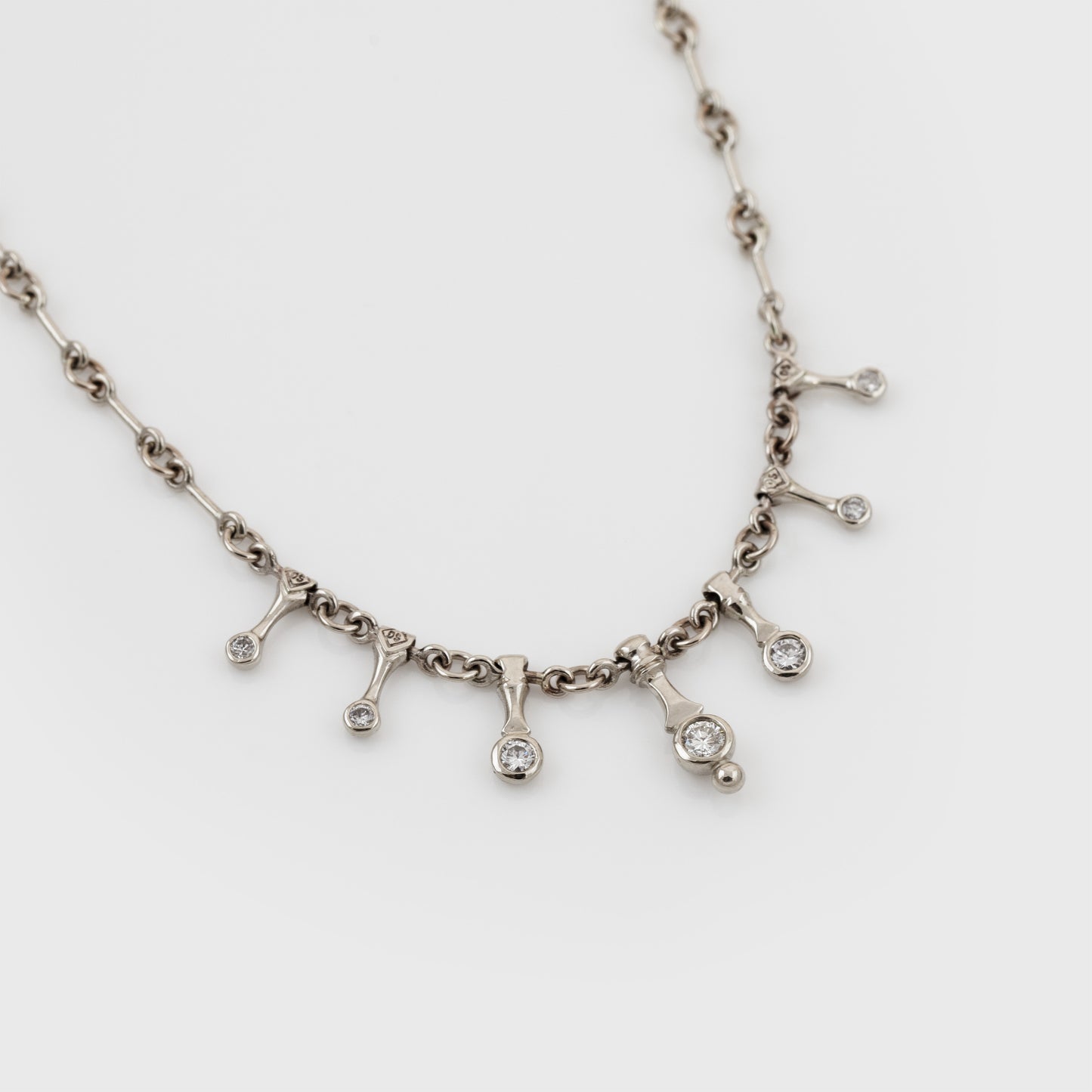 A silver-toned necklace with a chain and seven small dangling pendant charms, each set with a diamond, displayed on a light gray background.