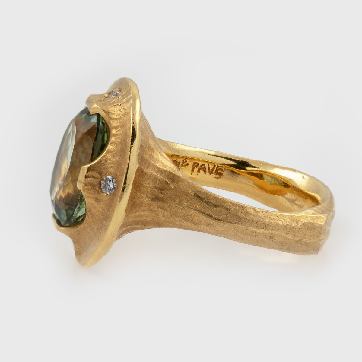 A textured yellow gold ring with a large oval green gemstone and a small round diamond accent, shown on a light gray background. The inside of the band is engraved with the word 'PAVE'.