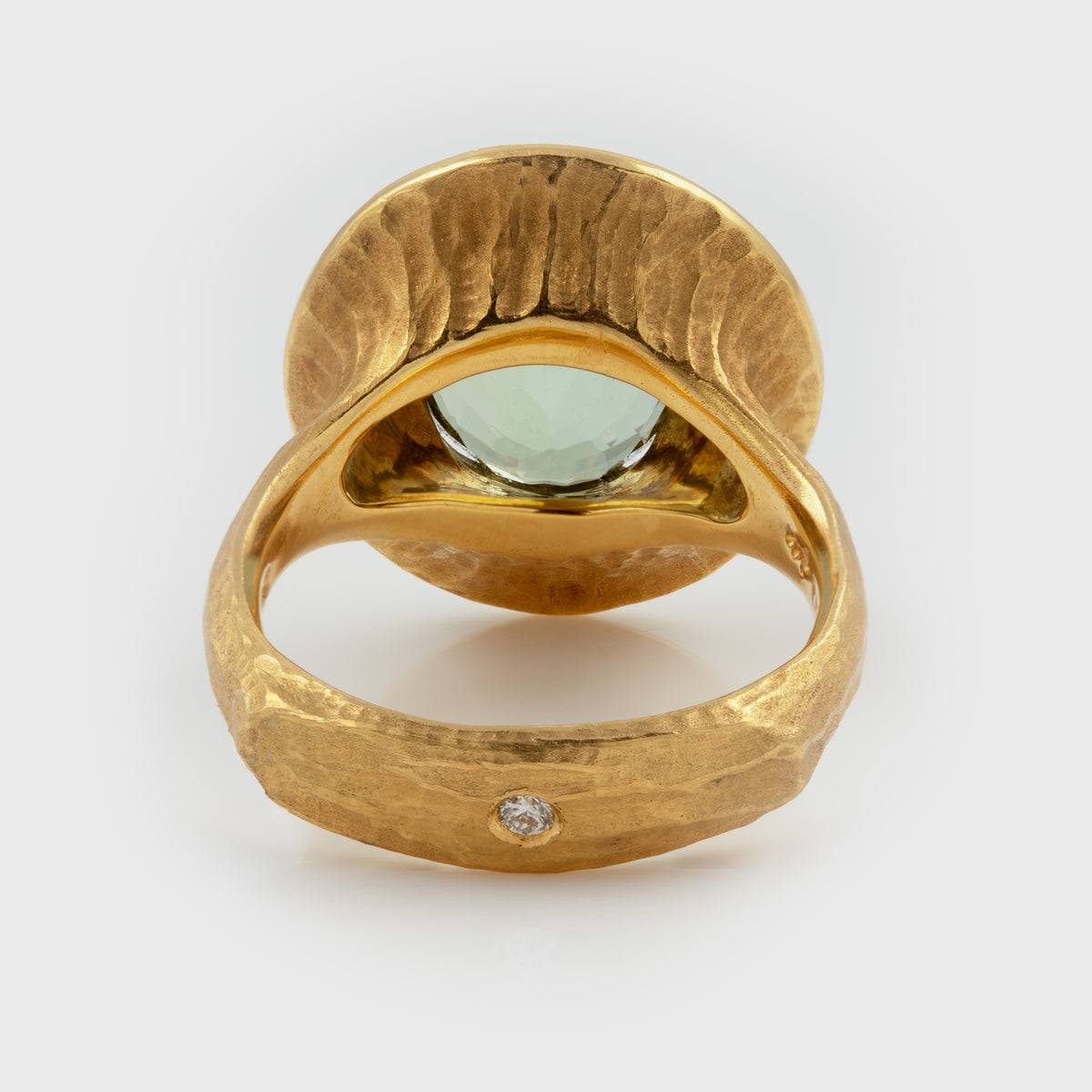 A textured yellow gold ring featuring a large oval light green gemstone in the center and a small round diamond set on the band, photographed on a white background.