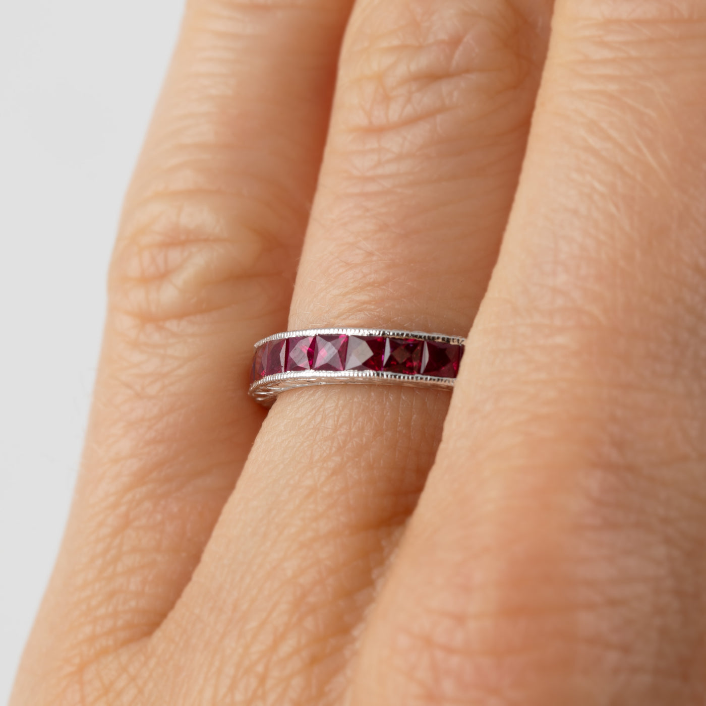 A close-up of a silver band ring with a row of faceted dark red gemstones, worn on a person's finger.