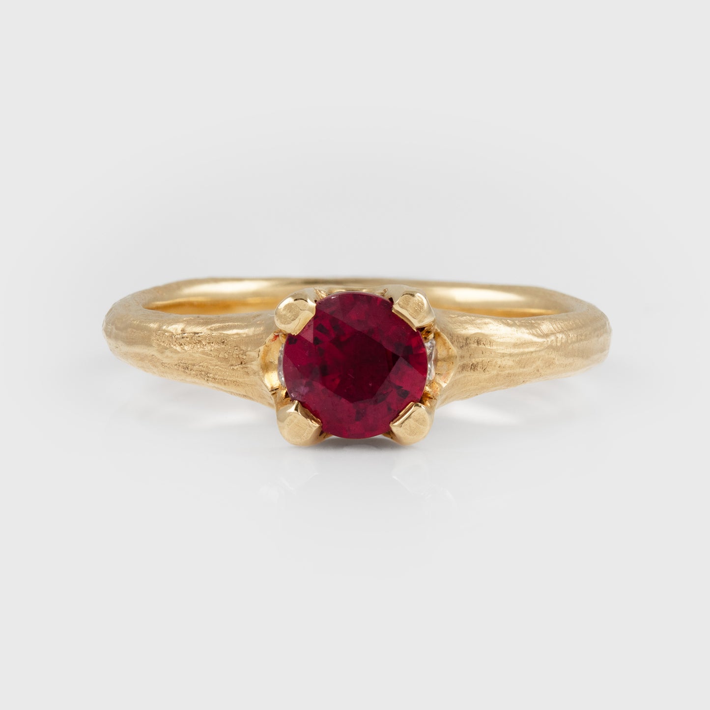 A gold ring with a textured band and a round-cut deep red gemstone in a prong setting, photographed on a light gray background.