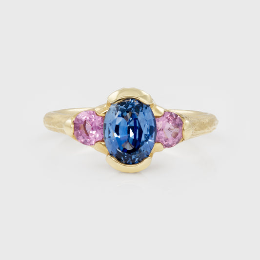 Gold ring with a blue gemstone and pink side stones on a light gray background