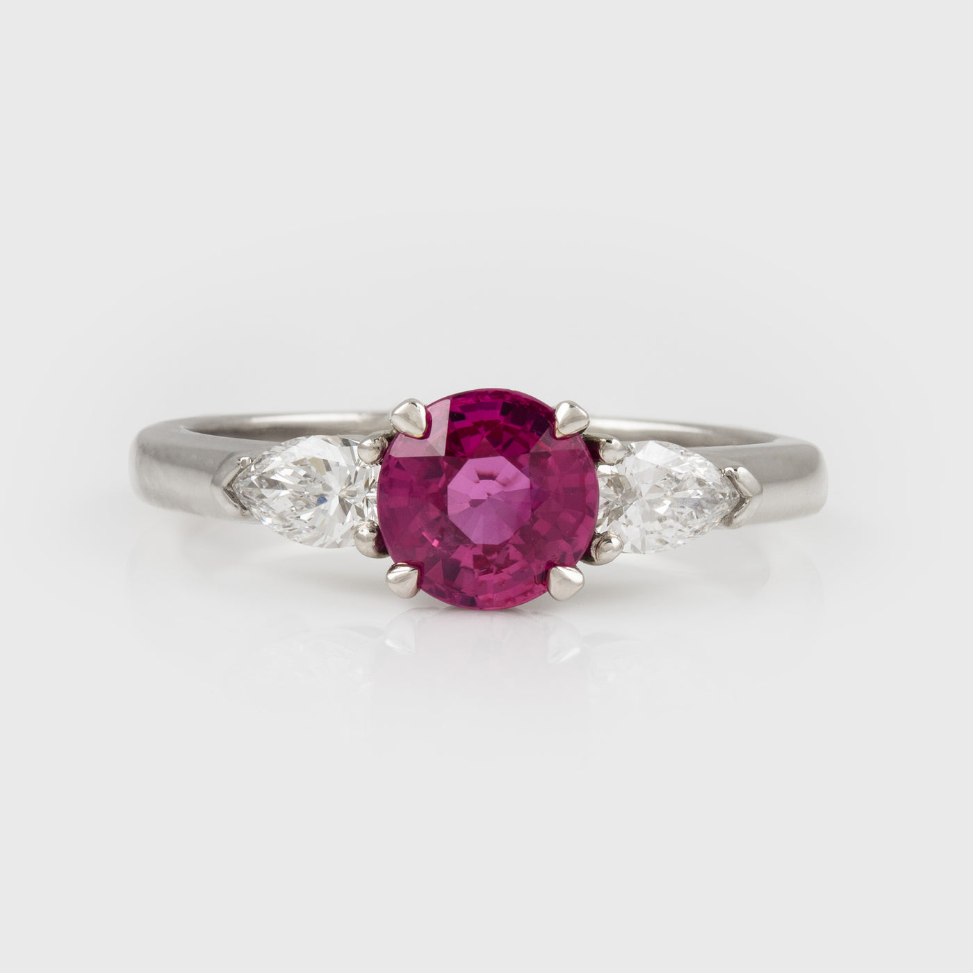 A platinum three-stone ring featuring a round-cut pinkish-red center ruby gemstone flanked by two marquise-cut clear side stones, set against a light gray background.