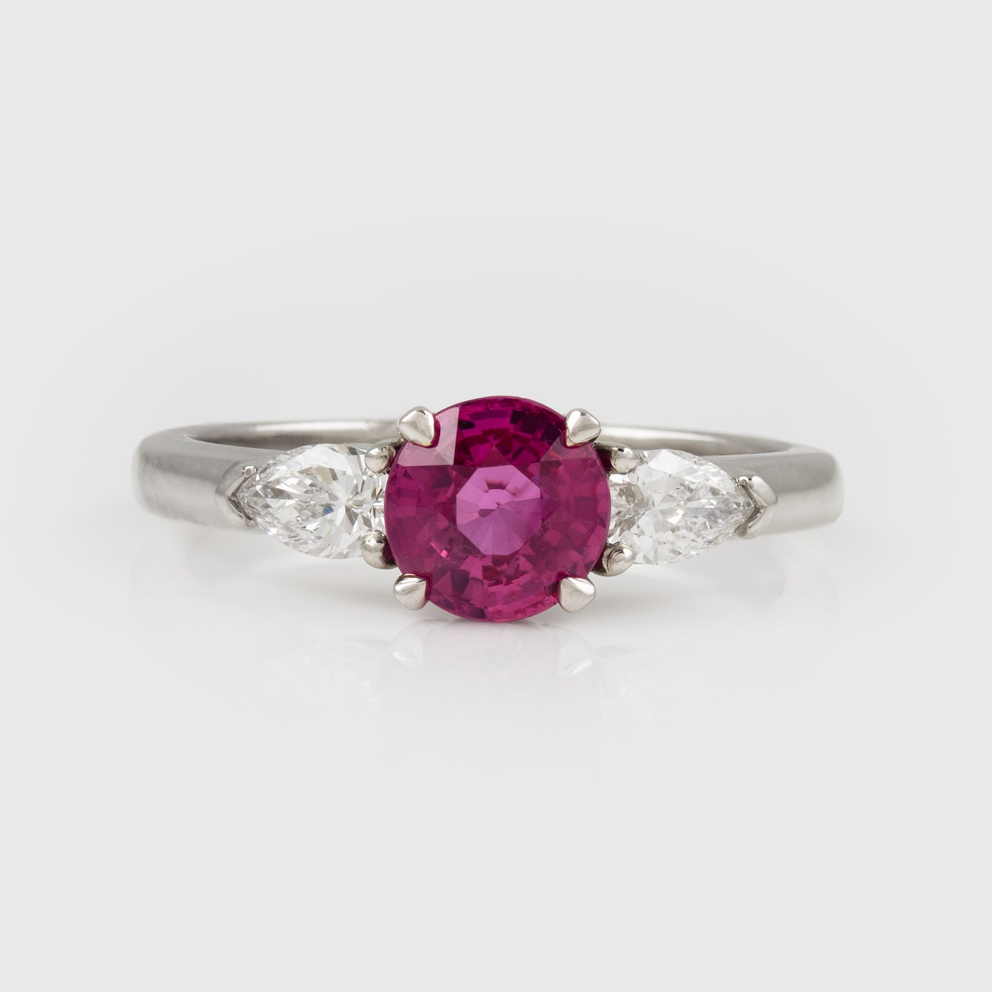 A platinum three-stone ring featuring a round-cut pinkish-red center ruby gemstone flanked by two marquise-cut clear side stones, set against a light gray background.