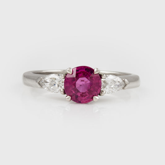 A platinum three-stone ring featuring a round-cut pinkish-red center ruby gemstone flanked by two marquise-cut clear side stones, set against a light gray background.