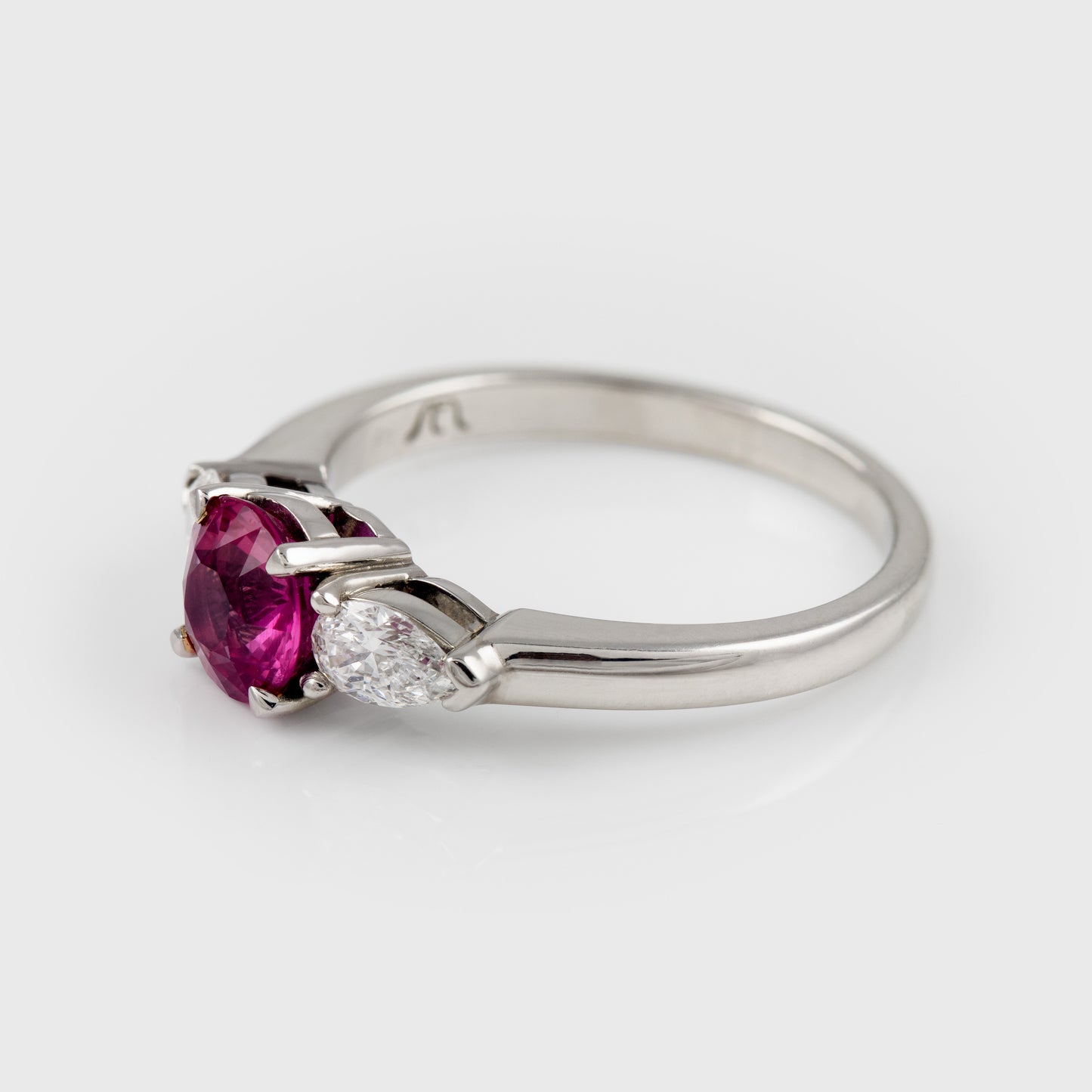 A silver ring featuring a round-cut pinkish-red ruby center stone and accent diamonds on a polished platinum band, set against a light gray background.