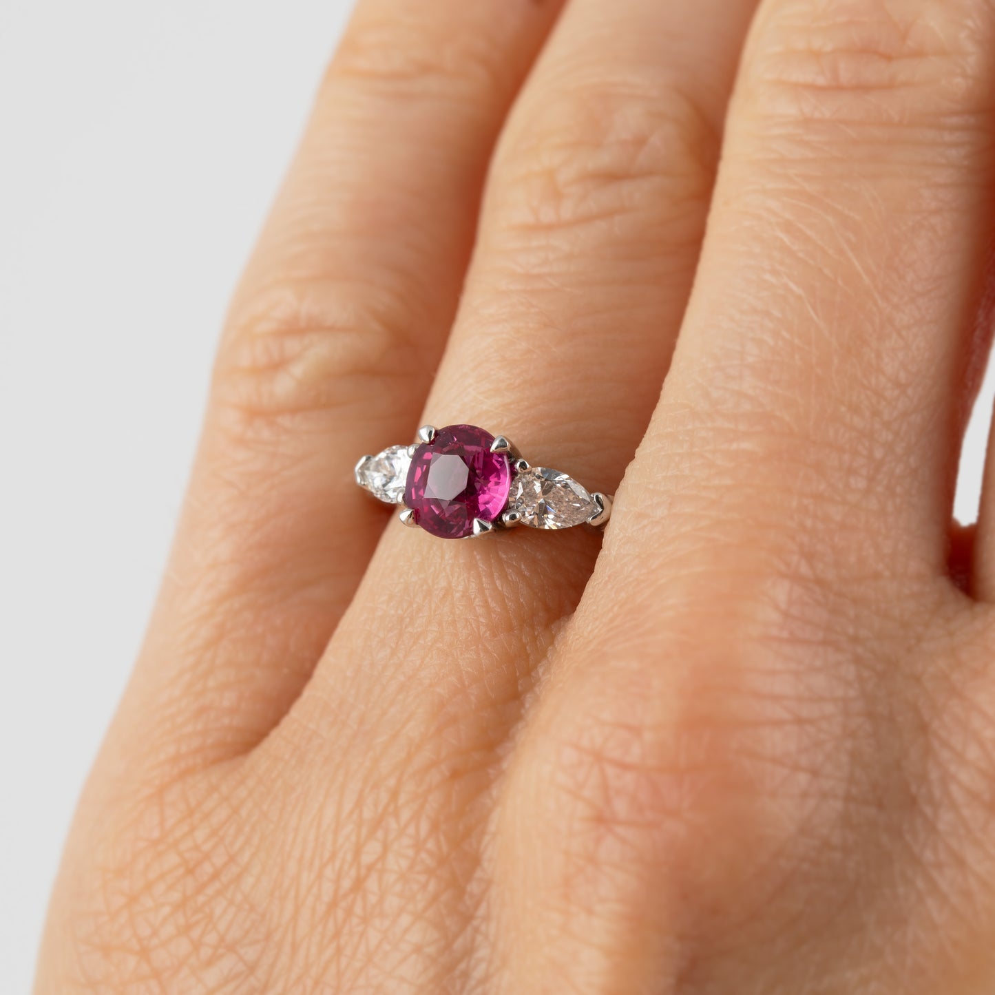 A close-up of a hand wearing a platinum ring with a central oval-cut pinkish red ruby gemstone and small diamond accent stones on either side.