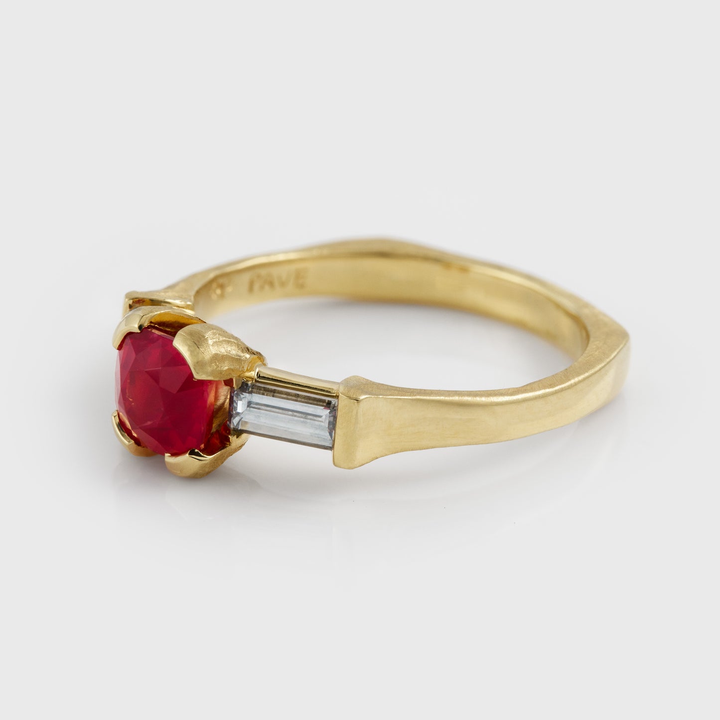 A yellow gold ring featuring a central red  o pink spinel gemstone flanked by two diamonds, set against a light gray background at an angled view