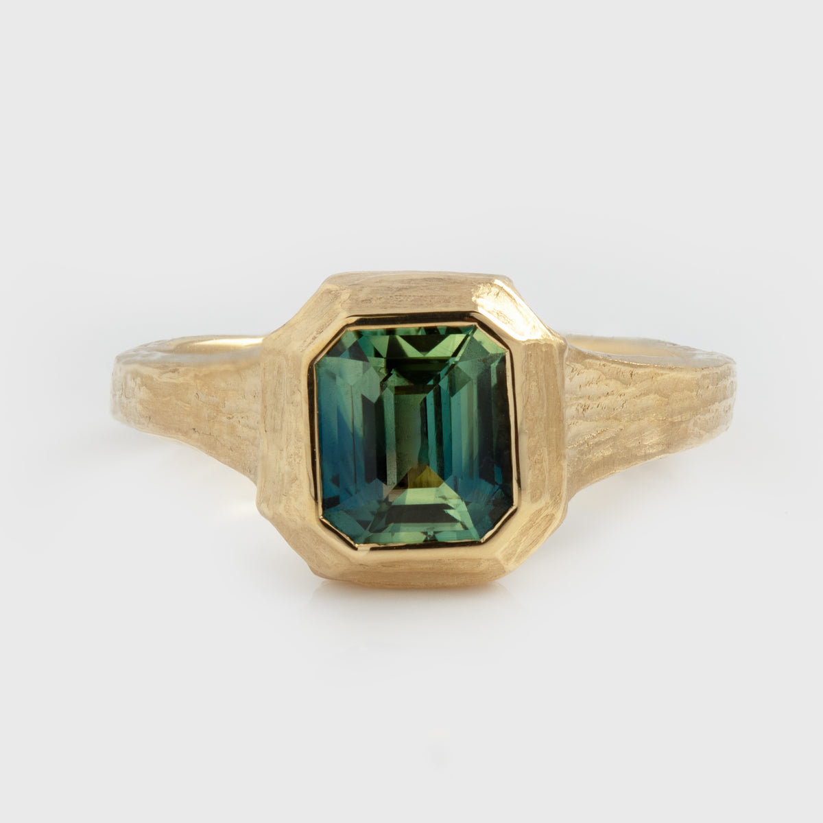 Yellow gold ring featuring a blue and green sapphire center stone with a textured band, photographed on a white background.