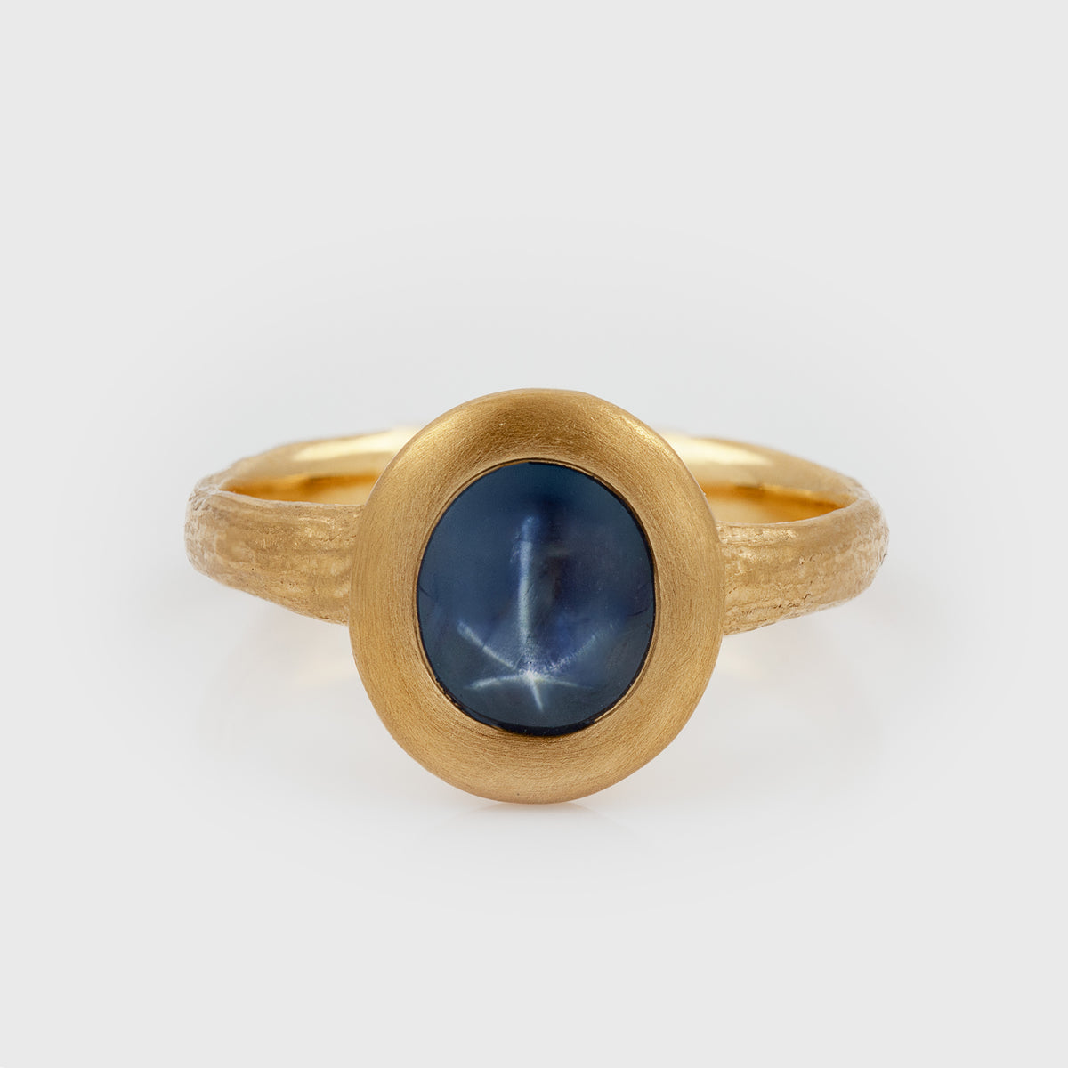Yellow gold ring featuring an oval blue Star Sapphire center stone with a textured band, photographed on a white background.
