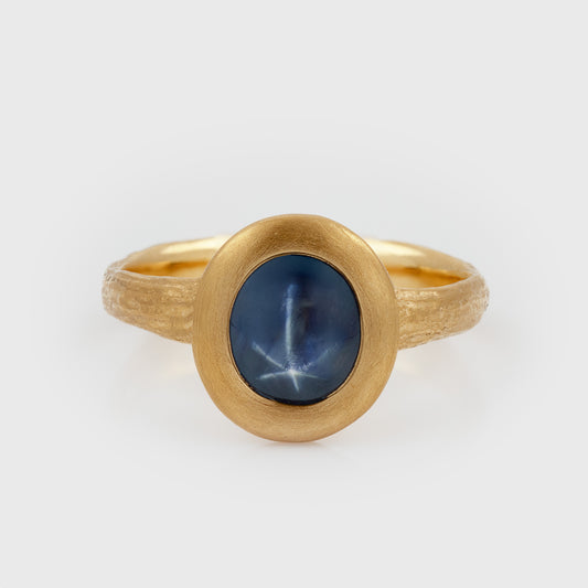 Yellow gold ring featuring an oval blue Star Sapphire center stone with a textured band, photographed on a white background.
