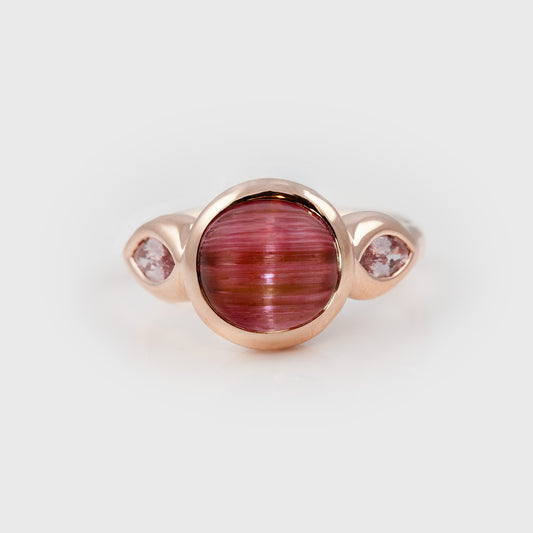 Ring featuring a pink cat’s eye center stone accented by pink sapphire, photographed on a white background.
