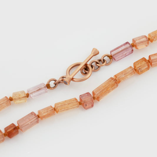 Beaded necklace featuring topaz beads in varying shades of orange and pink, accented with a gold toggle clasp, photographed on a white background.