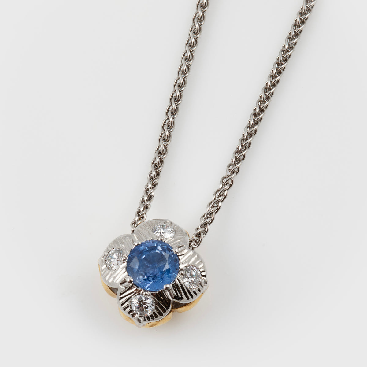 Flower pendant featuring blue sapphires and diamonds, suspended from a platinum chain, photographed on a white background.