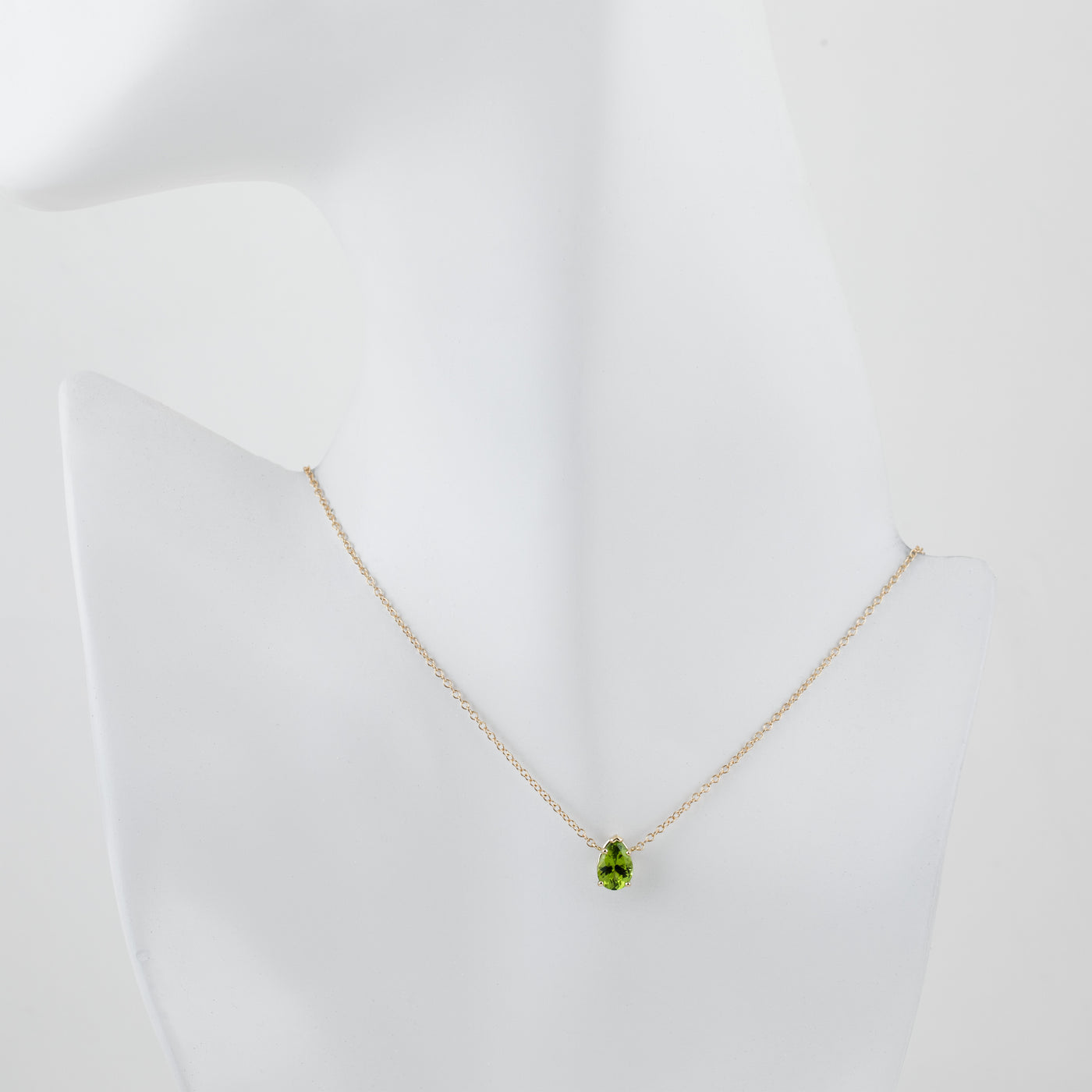 A delicate gold chain necklace with a green teardrop-shaped peridot gemstone pendant, displayed on a white mannequin bust against a clean white background.