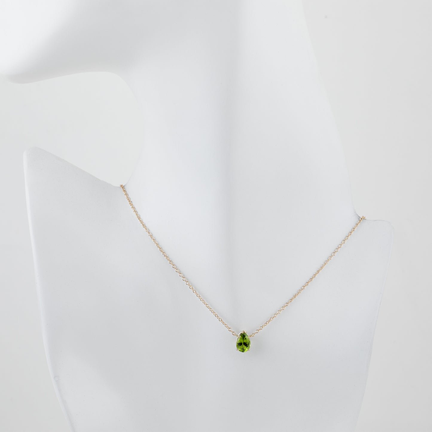 A delicate gold chain necklace with a green teardrop-shaped peridot gemstone pendant, displayed on a white mannequin bust against a clean white background.