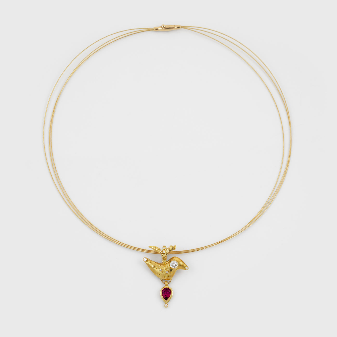 A delicate triple-strand gold necklace featuring a small bird-shaped pendant with a diamond and a dangling red teardrop gemstone, set against a plain light gray background.