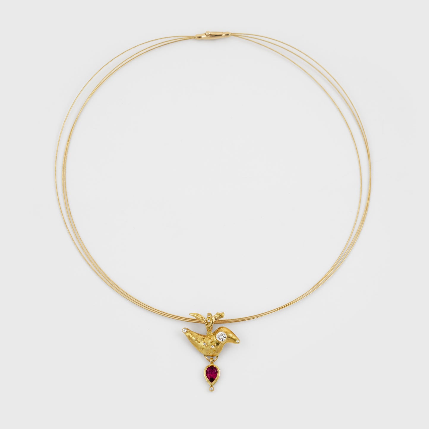 A delicate triple-strand gold necklace featuring a small bird-shaped pendant with a diamond and a dangling red teardrop gemstone, set against a plain light gray background.