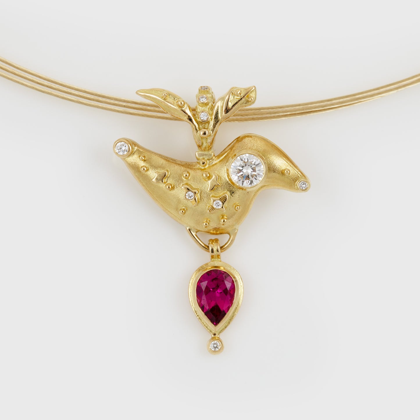 A gold bird pendant necklace featuring a gold bird body with a round diamond for its eye, more small diamonds, and a dangling teardrop-shaped deep pink gemstone, shown on a gold chain against a light gray background.