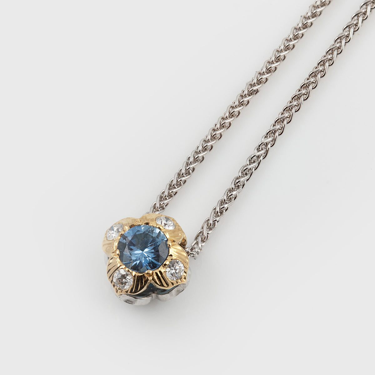 A flower pendant necklace featuring a round blue gemstone surrounded by small diamonds stones and textured gold-toned detailing, suspended from a silver-toned chain on a light gray background.
