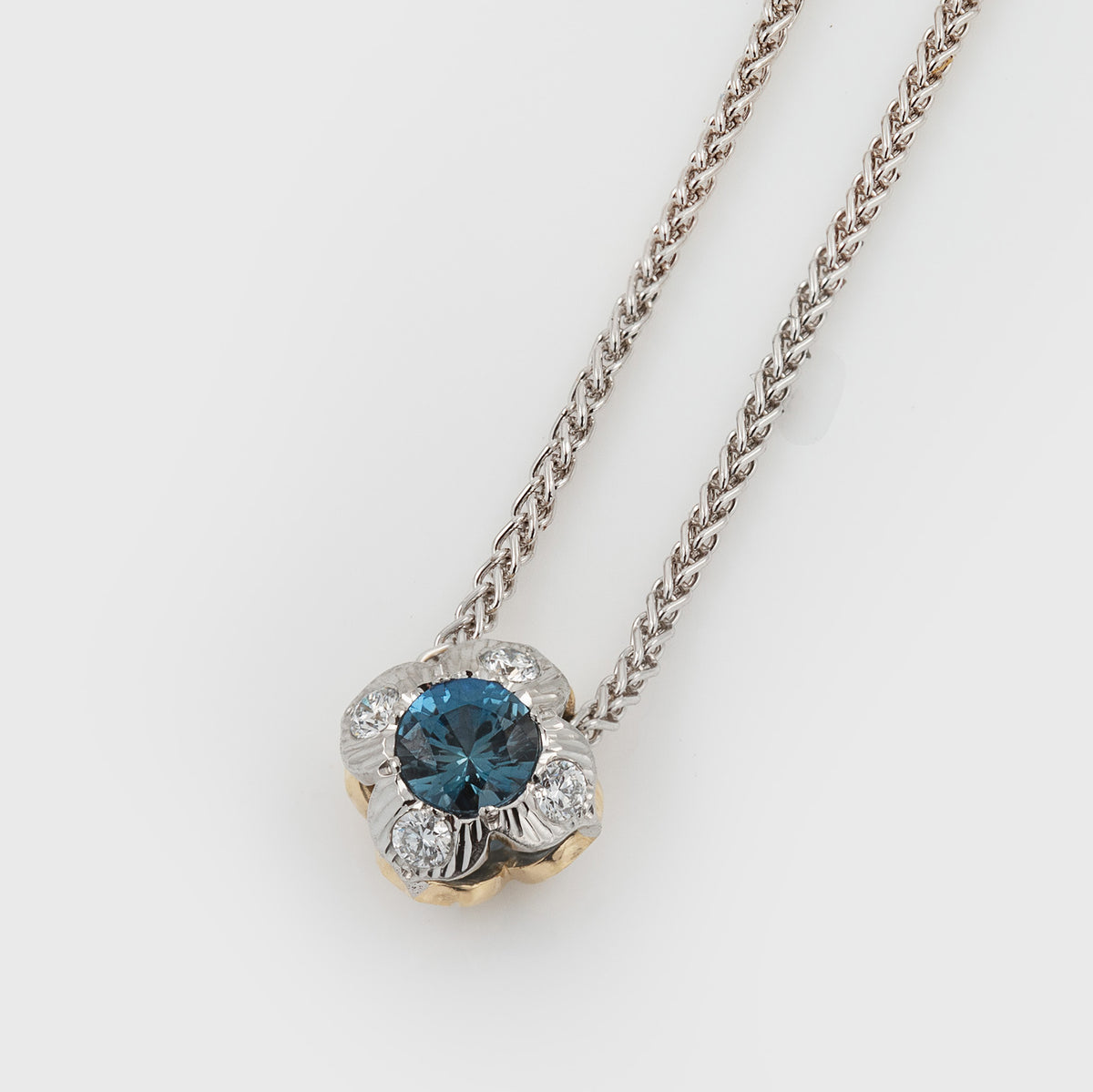 A flower pendant necklace featuring a round blue gemstone surrounded by small diamonds stones and textured platinum detailing, suspended from a silver-toned chain on a light gray background.
