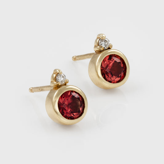 Pair of yellow gold round stud earrings with red spinel gemstones and accent diamonds on a white background