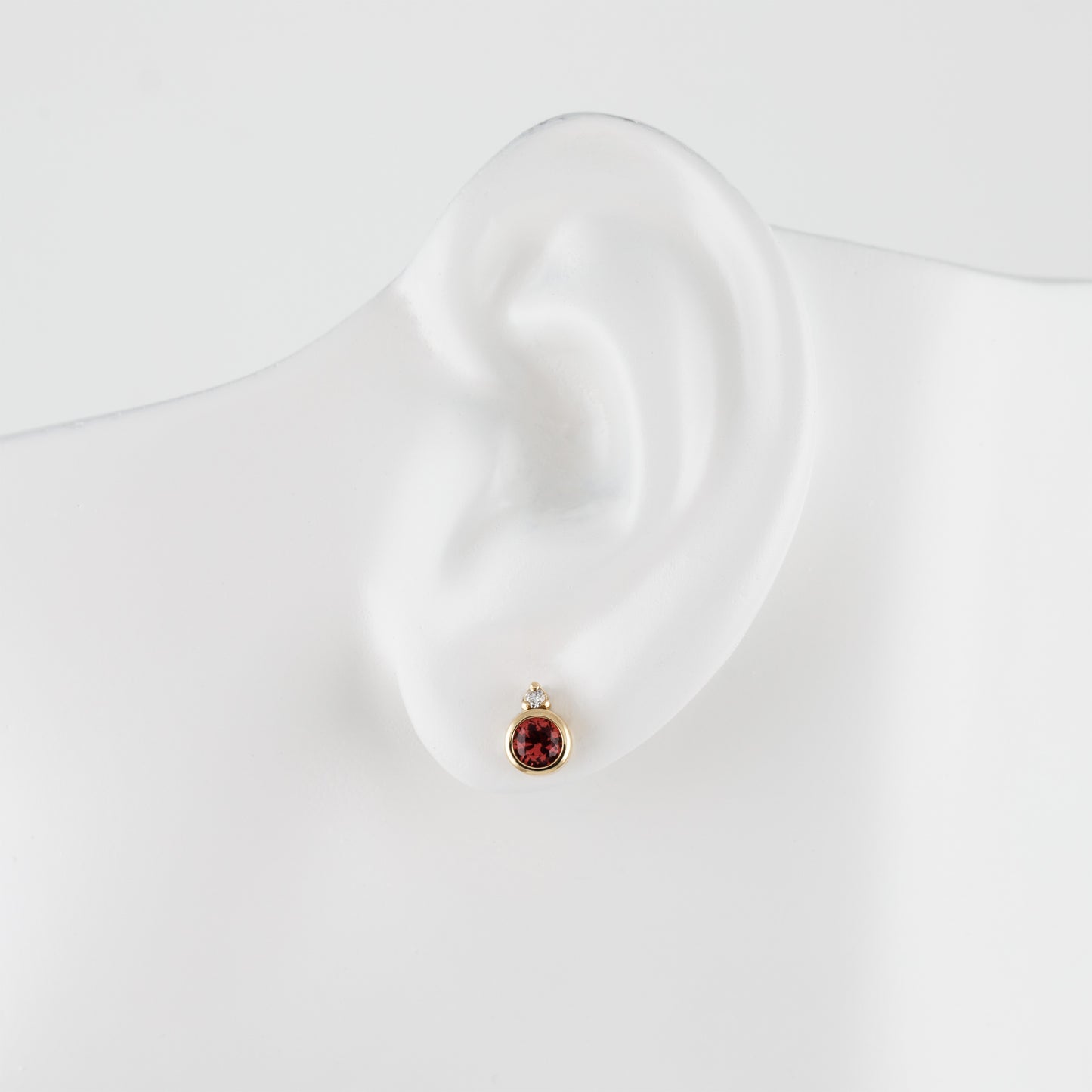 A single gold stud earring with a small red spinel gemstone displayed on a white ear mannequin against a clean white background.