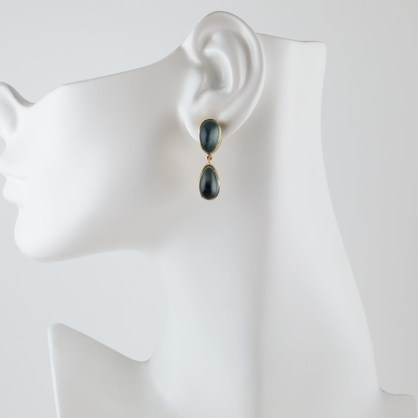 A pair of gold drop earrings with dark gray teardrop-shaped stones, displayed on a white mannequin head against a light gray background.