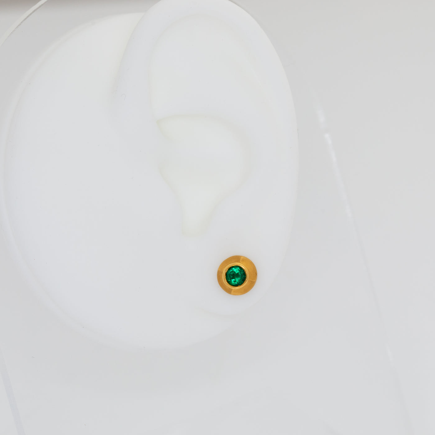 Close-up of a white ear mannequin with a green gemstone stud earring on a light gray background