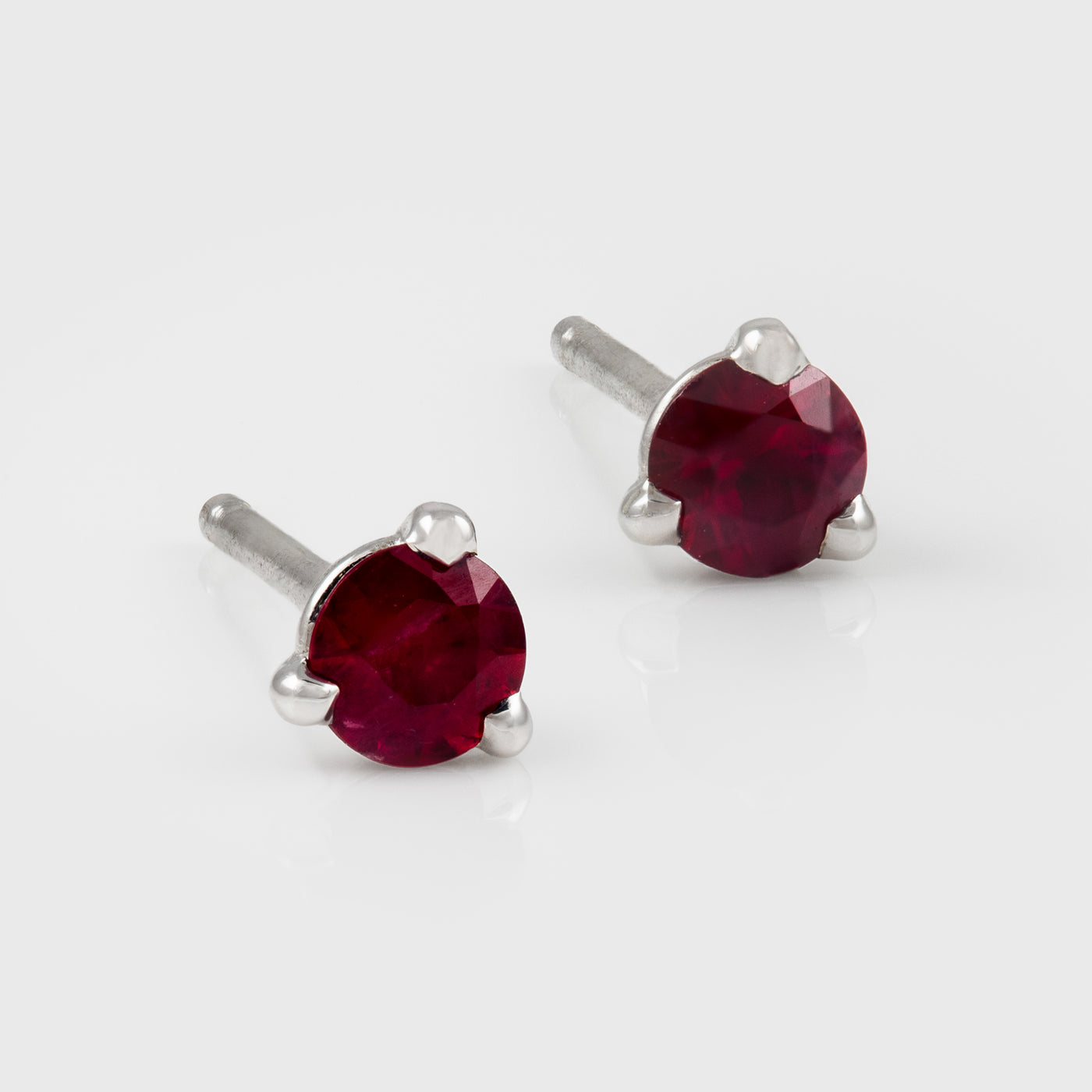 A pair of dark red ruby gemstone stud earrings in silver three-prong settings, shown on a clean light gray background.